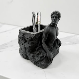 Peut inclure: Un organisateur de bureau noir mat composé d'un récipient carré à la texture rocheuse et irrégulière. Sur le côté est fixée une sculpture représentant un torse masculin aux cheveux bouclés, tourné sur le côté. Des stylos sont rangés dedans.