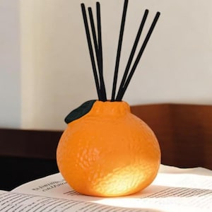 Op de afbeelding: Een oranje keramische diffuser in de vorm van een sinaasappel, met zwarte stokjes en een groene bladaccent. De diffuser staat op een open boek, met een vage achtergrond.