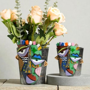 Pode incluir: Dois vasos decorativos com rosto, um grande e um pequeno, com designs abstratos coloridos. O vaso maior contém rosas cor de pêssego e vegetação. Os vasos têm um fundo cinza e são colocados em uma superfície de madeira.