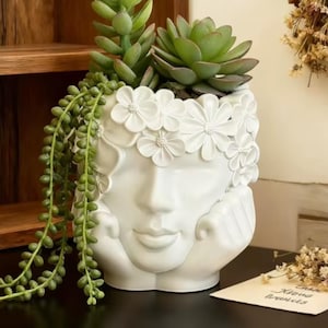Peut inclure: Un pot de fleurs en céramique blanche en forme de visage humain avec les mains sous le menton. La tête est ornée d'une couronne de fleurs blanches sculptées. Le pot contient une plante succulente verte et une plante retombante.