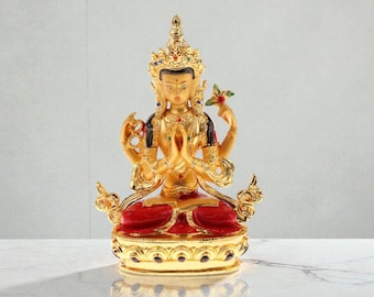 May include: A gold statue of a seated deity with multiple arms, wearing a jeweled crown and detailed embellishments. The figure sits on a red base with gold accents. The statue holds a flower and has hands in a prayer position.