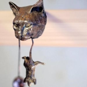 Könnte beinhalten: Eine Bronzeskulptur eines Katzenkopfes mit einer hängenden Katzenfigur. Der Katzenkopf hat geschlossene Augen und spitze Ohren. Die Skulptur ist an einem Holzbalken aufgehängt und erzeugt eine skurrile und künstlerische Darstellung.