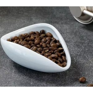Könnte beinhalten: Eine weiße, dreieckige Schale gefüllt mit gerösteten Kaffeebohnen. Die Bohnen haben eine satte braune Farbe und sehen frisch geröstet aus. Eine silberne Kaffeemühle ist im Hintergrund teilweise zu sehen.