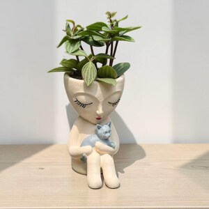 May include: A cream-colored ceramic planter shaped like a person with closed eyes and red lips, holding a small gray cat. The planter features a green plant with striped leaves growing from the top of the head, creating a cohesive decorative piece.