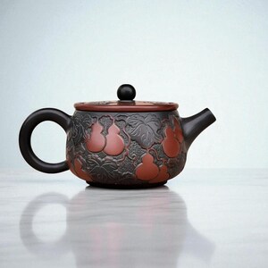 May include: A ceramic teapot with a black handle, spout, and lid. The body is dark grey with a pattern of red gourds and leaves. The lid has a black knob. The teapot sits on a white surface.