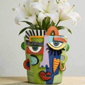 Könnte beinhalten: Eine dekorative Keramik-Gesichtsvase mit grünem Sockel, die farbenfrohe abstrakte Gesichtszüge aufweist. Die Vase enthält weiße Lilien. Das Design umfasst blaue Augen, rote Lippen sowie orangefarbene und blaue Akzente. Ein einzigartiges Wohnaccessoire.