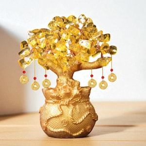 Op de afbeelding: Een gouden decoratieve boom met gele kristallen bladeren en een pot met textuur. De boom heeft hangende gouden munten met rode kralen. De boom staat op een houten oppervlak.