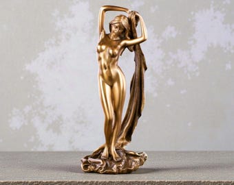 May include: A golden statue of a nude woman with her arms raised, holding a draped cloth. The statue is on a textured base and has a smooth, polished finish. The background is a neutral gray.