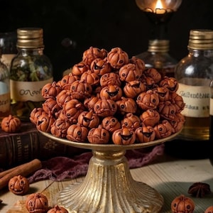 May include: A large pile of small, carved brown pumpkin-shaped beads with carved facial expressions, resting on a gold-colored pedestal stand. The pumpkins feature detailed ridges and cut-out eyes and mouths, creating a textured, uniform appearance.