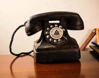 May include: An antique black rotary dial telephone with a handset and a coiled cord. The phone has a circular dial with numbers and a central emblem. It sits on a wooden surface, showcasing a vintage aesthetic.