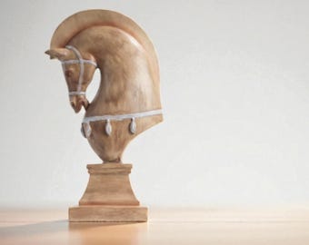 Puede incluir: Escultura de cabeza de caballo marrón claro con detalles blancos. La escultura está montada sobre una base rectangular. La cabeza del caballo presenta una crin, una brida y elementos decorativos. La escultura es de madera y es un objeto decorativo.