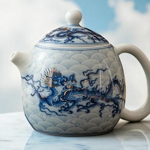 Op de afbeelding: Een witte keramische theepot met een blauw drakenontwerp. De theepot heeft een gebogen handvat, een tuit en een deksel met een kleine knop. De draak is gedetailleerd en gestileerd afgebeeld, met gouden accenten.