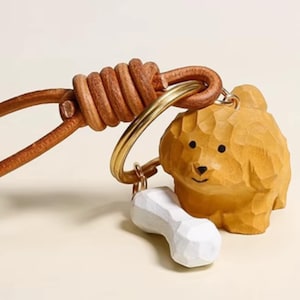 May include: A wooden dog keychain with a white bone charm. The dog is carved from light brown wood and has a smiling face. The keychain includes a gold-colored ring and a brown leather strap.
