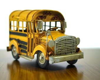 May include: A miniature yellow school bus with black stripes, a silver grill, and small wheels. The vintage-style bus has a detailed design, including windows and mirrors, and sits on a wooden surface.