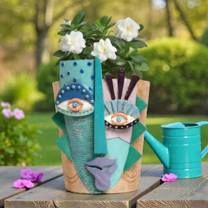 Puede incluir: Una maceta decorativa de cerámica con forma de rostro, con nariz y detalles turquesa, diseños de ojos y boca morada. La maceta contiene una planta con flores blancas. Una regadera turquesa está cerca.