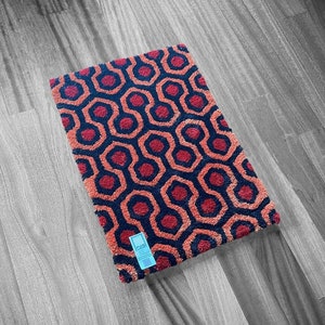 Op de afbeelding: Een rechthoekige deurmat met een herhalend geometrisch patroon in tinten rood, marineblauw en oranje. Het ontwerp bevat onderling verbonden zeshoekige vormen. Een klein blauw label is aan de mat bevestigd. De deurmat ligt op een houten vloer.