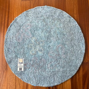 May include: A round, light blue rug with a textured surface. The rug has a faded floral design in shades of pink and yellow. A rectangular tag with text and a QR code is attached to the rug.