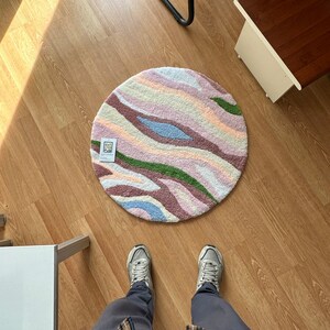 May include: A round rug with a colorful, abstract design. The rug features a pattern of wavy stripes in shades of pink, green, blue, and cream. The rug is placed on a wooden floor.