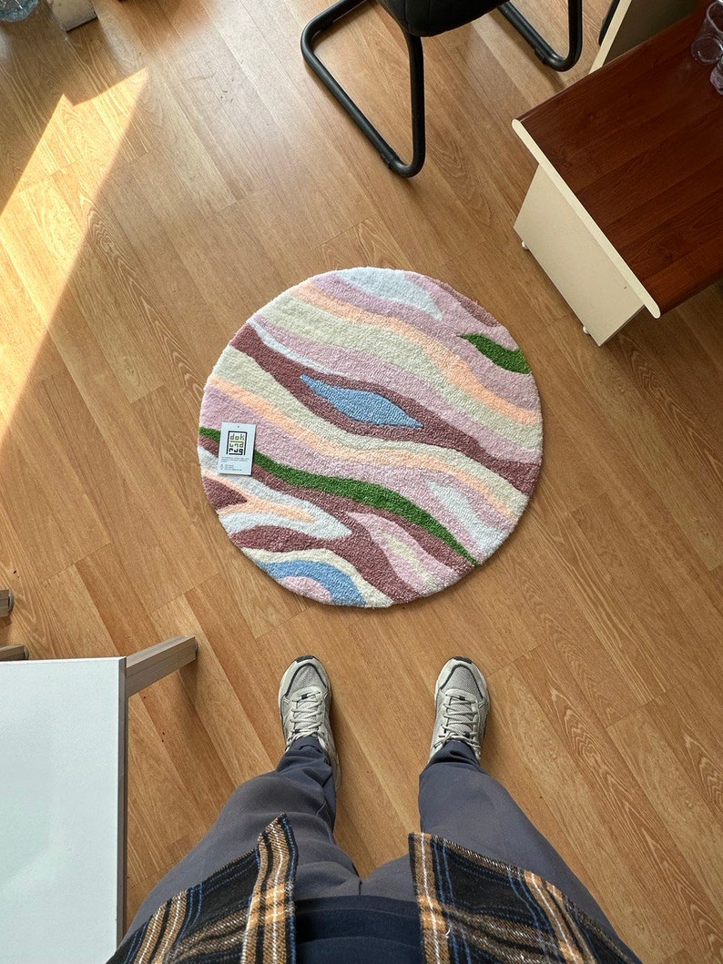 May include: A round rug with a colorful, abstract design. The rug features a pattern of wavy stripes in shades of pink, green, blue, and cream. The rug is placed on a wooden floor.