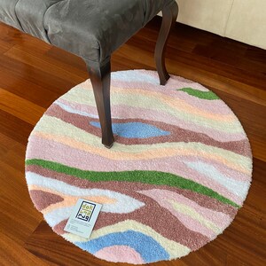 May include: A round, tufted rug with a colorful, abstract design. The rug features wavy stripes in shades of pink, brown, green, blue, and white. A gray bench with dark wooden legs is partially visible above the rug. A business card with text is on the rug.