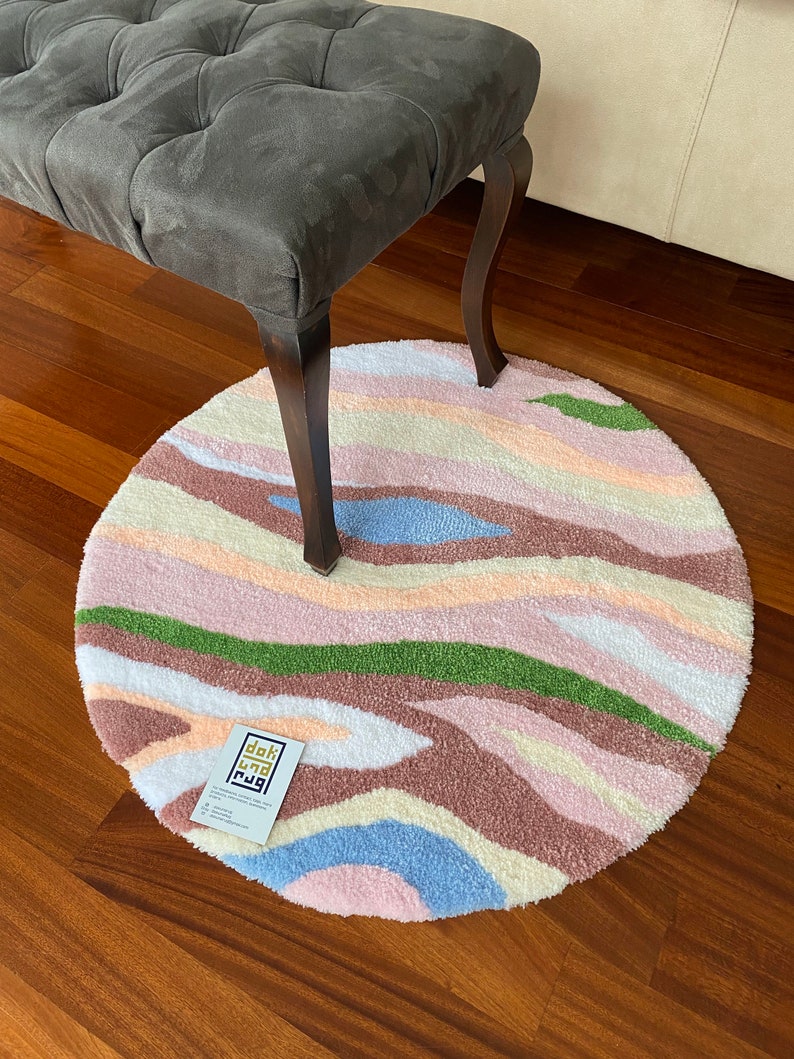 May include: A round, tufted rug with a colorful, abstract design. The rug features wavy stripes in shades of pink, brown, green, blue, and white. A gray bench with dark wooden legs is partially visible above the rug. A business card with text is on the rug.