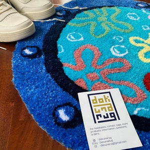 May include: A round, blue and turquoise rug with a cartoon design, featuring a white and cream-colored sneaker. The rug has a textured surface and a dark blue border. A business card with contact information is also visible.