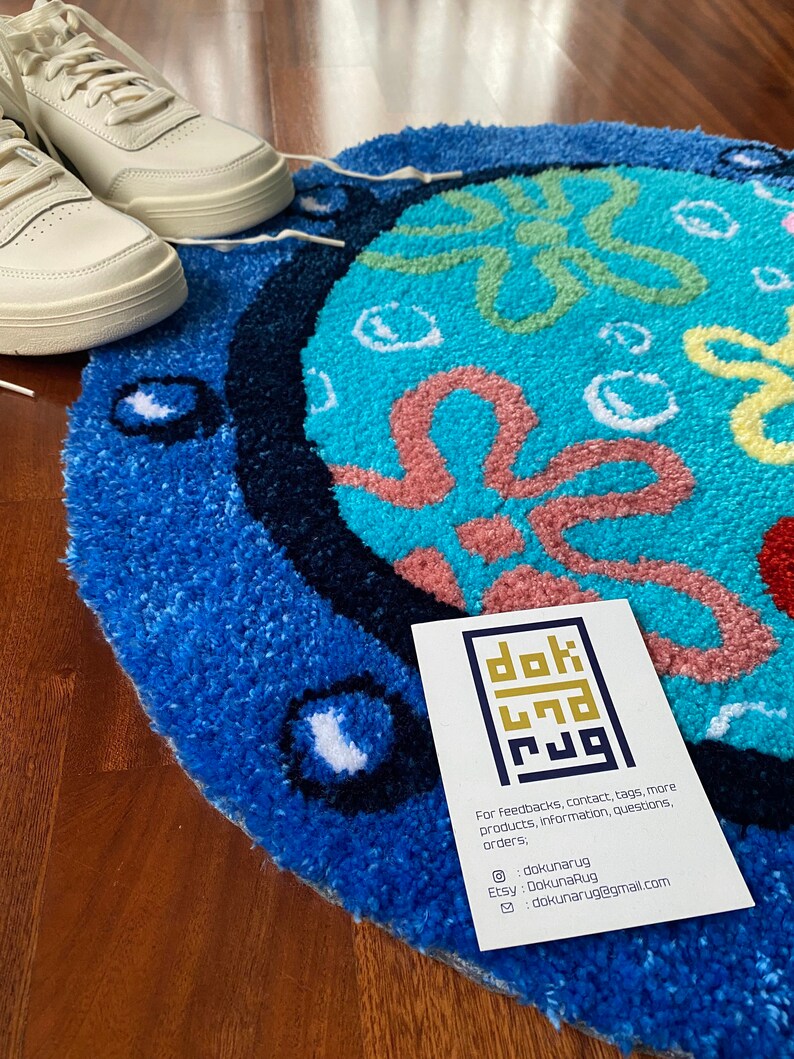 May include: A round, blue and turquoise rug with a cartoon design, featuring a white and cream-colored sneaker. The rug has a textured surface and a dark blue border. A business card with contact information is also visible.