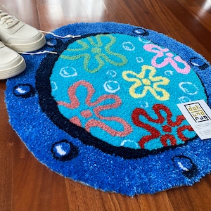 May include: A round, blue rug with a cartoon floral design in shades of red, yellow, and pink. The rug has a dark blue border and white accents. White sneakers are visible in the background.