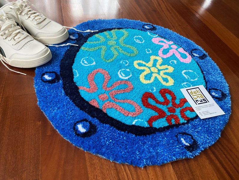 May include: A round, blue rug with a cartoon floral design in shades of red, yellow, and pink. The rug has a dark blue border and white accents. White sneakers are visible in the background.
