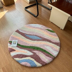 May include: A round rug with a colorful, abstract design. The rug features wavy stripes in shades of pink, purple, green, blue, and cream. The rug has a soft, textured appearance. A tag with text is attached to the rug.