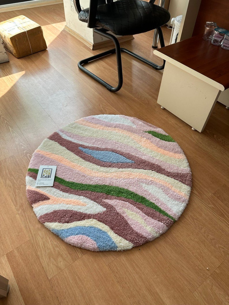 May include: A round rug with a colorful, abstract design. The rug features wavy stripes in shades of pink, purple, green, blue, and cream. The rug has a soft, textured appearance. A tag with text is attached to the rug.