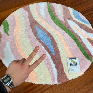 May include: A round rug with a colorful, abstract design. The rug features vertical stripes in shades of pink, beige, green, blue, and brown. A small tag with text is visible on the rug.