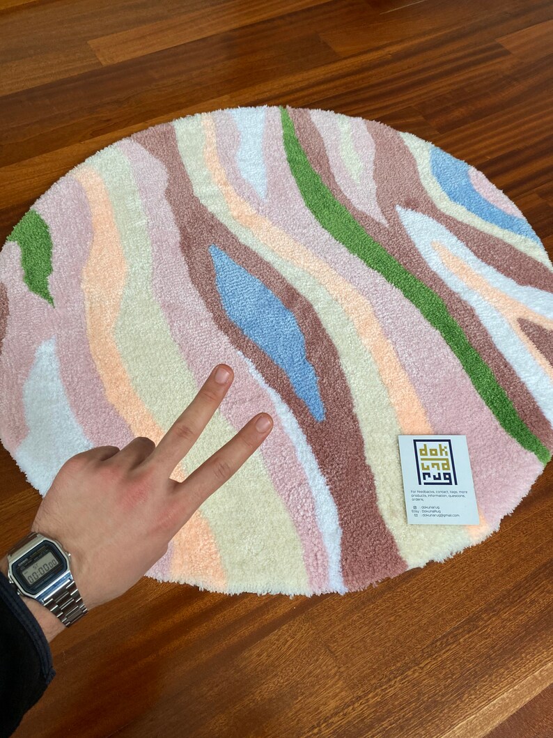 May include: A round rug with a colorful, abstract design. The rug features vertical stripes in shades of pink, beige, green, blue, and brown. A small tag with text is visible on the rug.