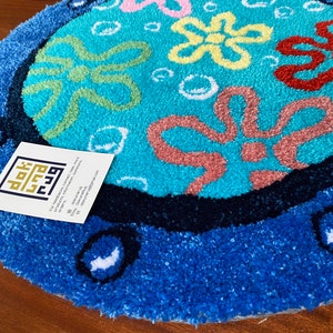 May include: A round, blue rug with a cartoon design. The rug features a light blue center with colorful flower-like shapes and white bubbles. The border is a darker blue, with white accents. The rug is on a wooden surface.