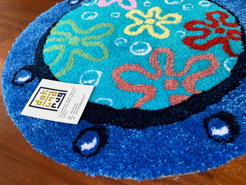 May include: A round, blue rug with a cartoon design. The rug features a light blue center with colorful flower-like shapes and white bubbles. The border is a darker blue, with white accents. The rug is on a wooden surface.