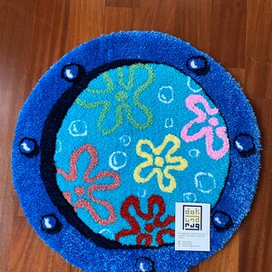 May include: A round, blue rug with a cartoon flower design. The rug features a blue border with bubble-like accents and a central design of colorful flowers on a light blue background. The rug has a textured, plush appearance.