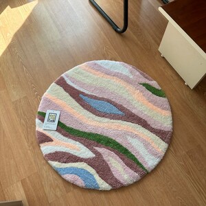 May include: A round, tufted rug with a wavy pattern in pastel colors. The rug features stripes of pink, purple, green, blue, and cream. The rug is on a wooden floor. A tag with text is attached to the rug.
