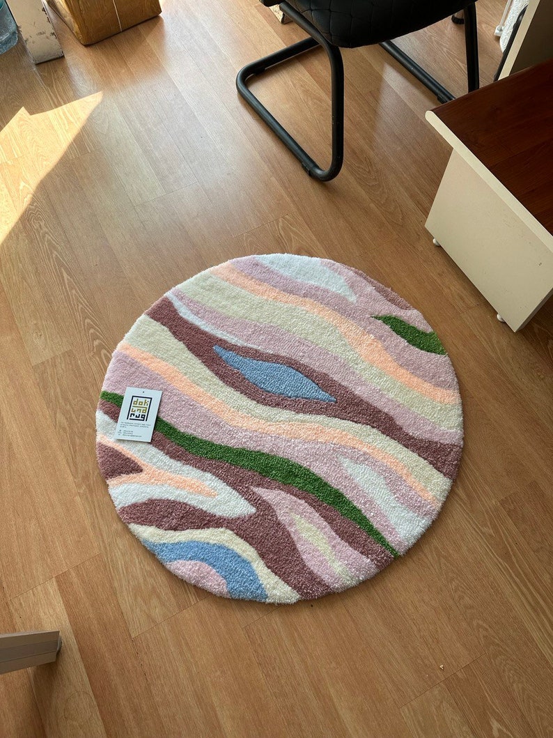 May include: A round, tufted rug with a wavy pattern in pastel colors. The rug features stripes of pink, purple, green, blue, and cream. The rug is on a wooden floor. A tag with text is attached to the rug.