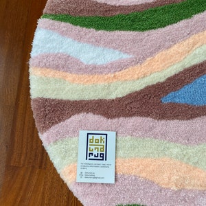 May include: A round, textured rug with a landscape design. The rug features horizontal bands of color including brown, green, pink, white, and blue. A business card with the text "dok rug" is on the rug.