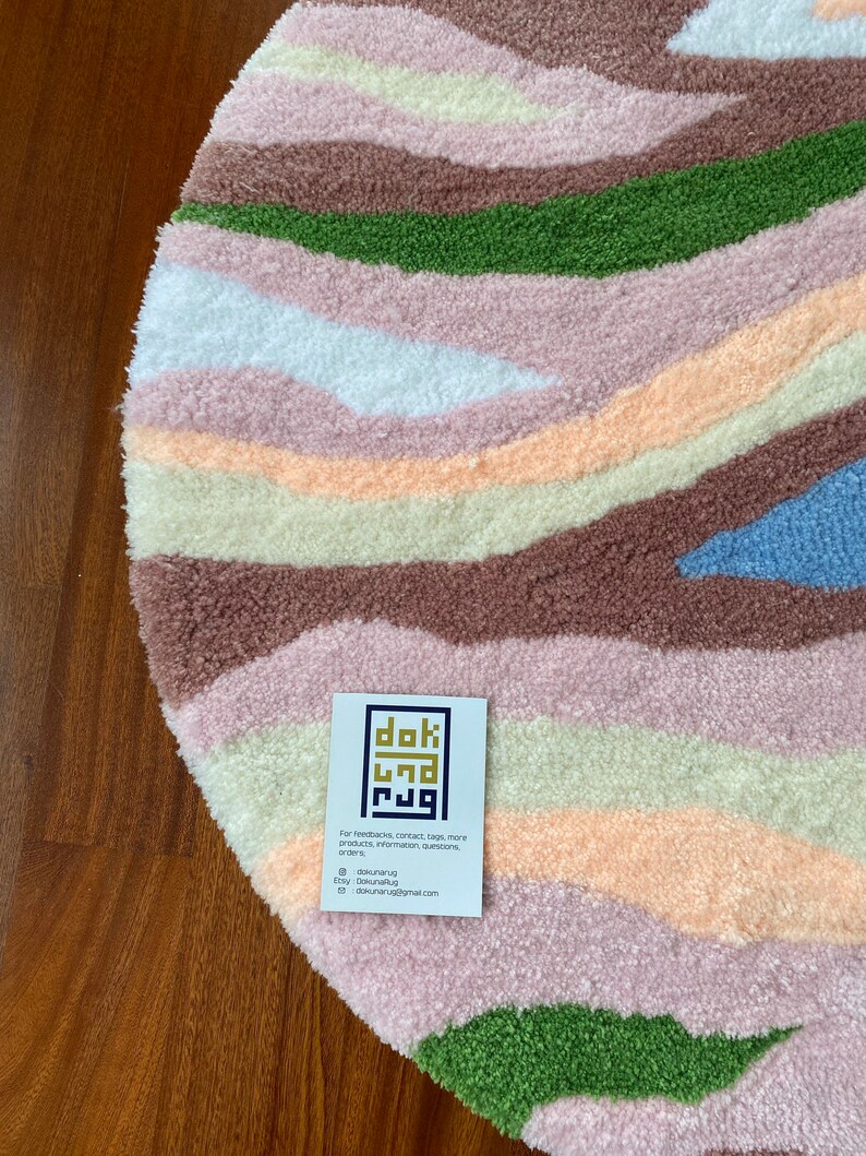May include: A round, textured rug with a landscape design. The rug features horizontal bands of color including brown, green, pink, white, and blue. A business card with the text "dok rug" is on the rug.