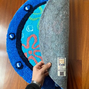 May include: A half-circle rug with a blue border and a cartoon design in shades of blue, teal, and coral. The rug's backing is gray. The rug is displayed on a wooden surface.