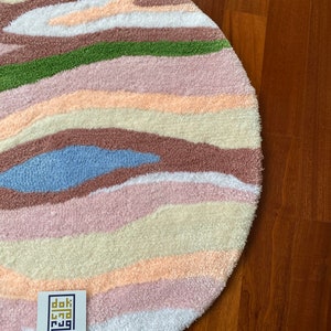 May include: A round, tufted rug with a colorful, abstract design. The rug features horizontal bands of various colors, including white, pink, green, brown, and blue. A small white tag with black text is visible in the lower left corner.