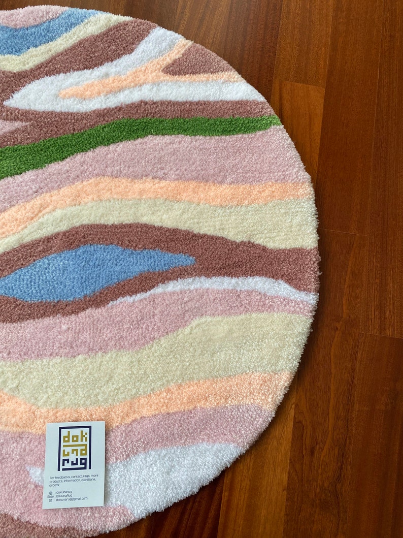 May include: A round, tufted rug with a colorful, abstract design. The rug features horizontal bands of various colors, including white, pink, green, brown, and blue. A small white tag with black text is visible in the lower left corner.