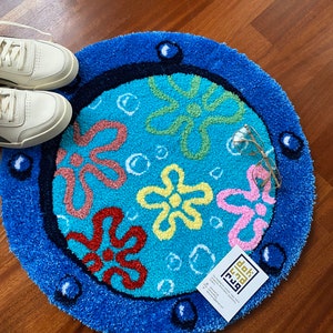May include: A round, blue rug with a cartoon flower design. The rug features a light blue center with colorful flowers and bubbles, surrounded by a dark blue border. A pair of white sneakers and eyeglasses are next to the rug.