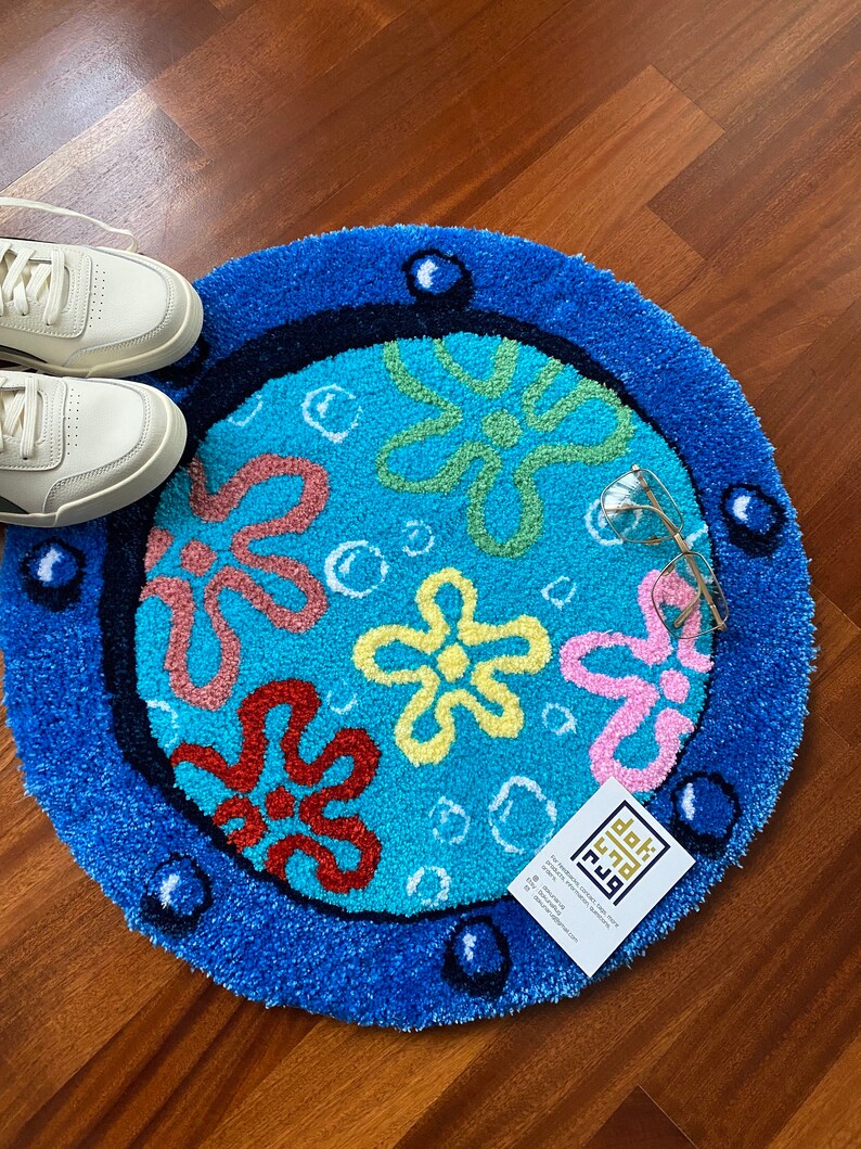May include: A round, blue rug with a cartoon flower design. The rug features a light blue center with colorful flowers and bubbles, surrounded by a dark blue border. A pair of white sneakers and eyeglasses are next to the rug.