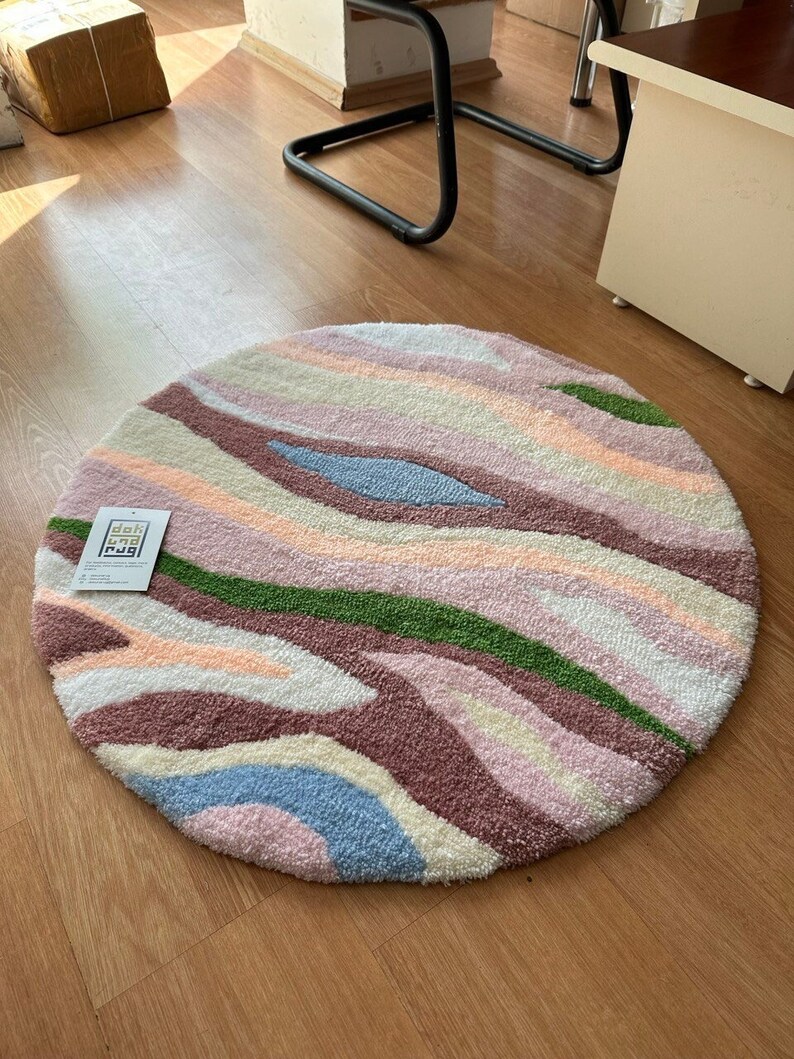 May include: A round rug with a colorful, abstract design. The rug features a pattern of wavy stripes in shades of pink, purple, green, blue, and cream. The rug has a soft, textured appearance and a label in the corner.