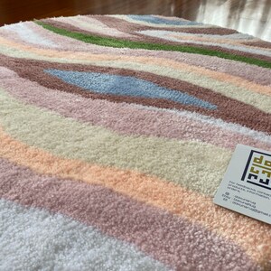 May include: A round rug with a wavy pattern in pastel colors. The rug features stripes of white, peach, pink, brown, green, and blue. The rug has a soft, textured appearance. A tag with the text "dok rug" is in the bottom right corner.