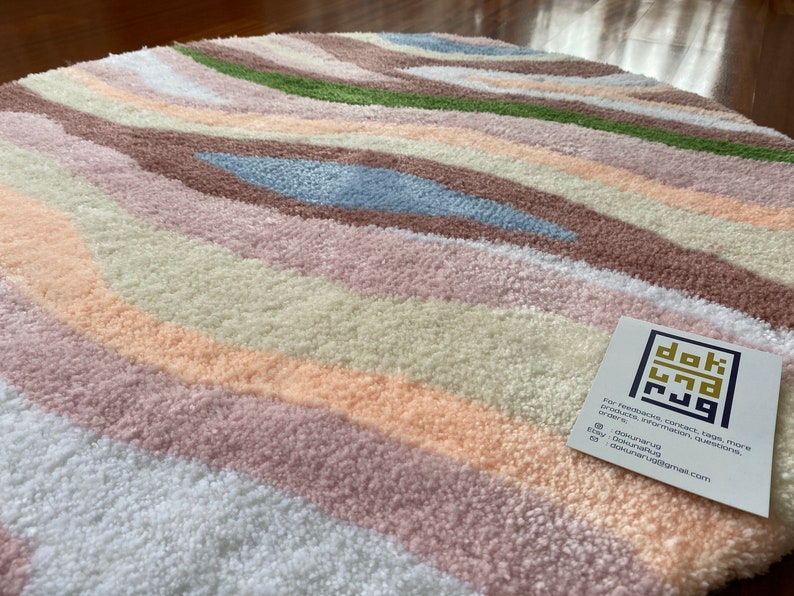 May include: A round rug with a wavy pattern in pastel colors. The rug features stripes of white, peach, pink, brown, green, and blue. The rug has a soft, textured appearance. A tag with the text "dok rug" is in the bottom right corner.