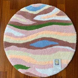 May include: A round, tufted rug with a wavy, abstract design. The rug features bands of color including light blue, brown, green, pink, and cream. The rug is made of a soft, textured material and is placed on a wooden floor.