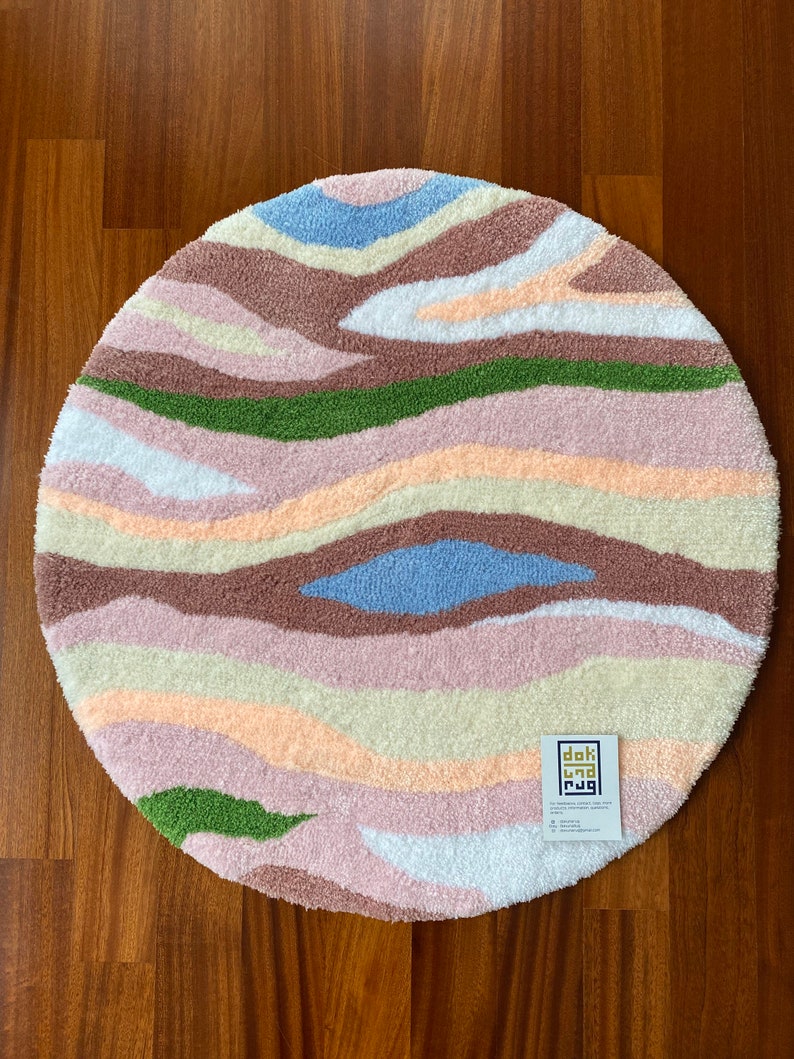May include: A round, tufted rug with a wavy, abstract design. The rug features bands of color including light blue, brown, green, pink, and cream. The rug is made of a soft, textured material and is placed on a wooden floor.